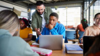 Male teacher helping a young college student with his work on a laptop while sitting with classmates at a table in a school cafeteria.