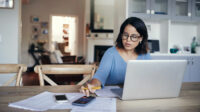 Shot of a young woman using a laptop and calculator while working from home