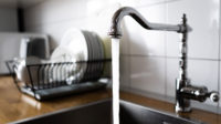 Water flowing out of a kitchen stainless steel tap into the sink. Wasting water by leaving a chrome faucet tap running. Water misuse in domestic duties and activities. Overusing household water.