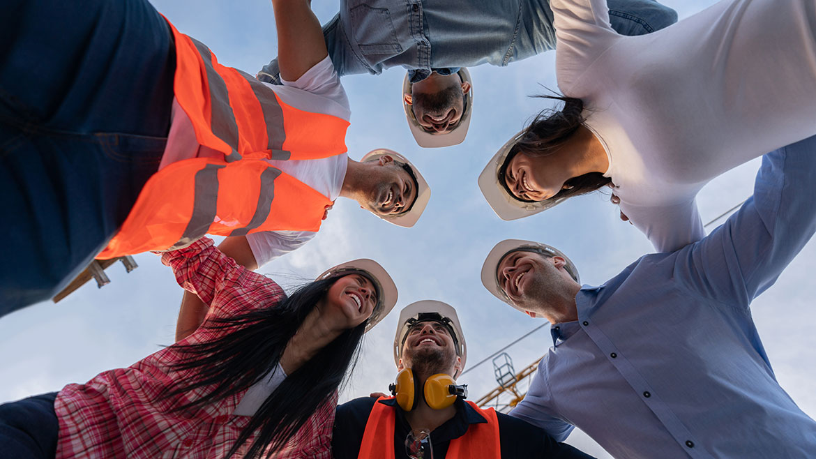 Happy team of architects hugging in a circle at a construction site smiling and wearing helmets.