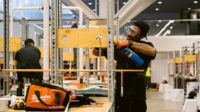 An apprentice working in a workshop, the back of another apprentice in the background.