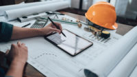 Engineer working with drawings inspection on a tablet in the office along with a calculator, triangle ruler, safety glasses, compass, vernier caliper on the blueprint.