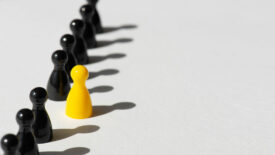 Black chess pieces in a line behind one yellow chess piece over white background.