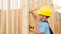 A young boy uses a measuring tape and pretends to be a construction worker. He is wearing a hard hat, suspenders, and a tool belt while measuring on a stud of a house being built in Utah, USA.