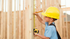 A young boy uses a measuring tape and pretends to be a construction worker. He is wearing a hard hat, suspenders, and a tool belt while measuring on a stud of a house being built in Utah, USA.