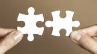 Hand holding two white puzzle pieces with a brown background.