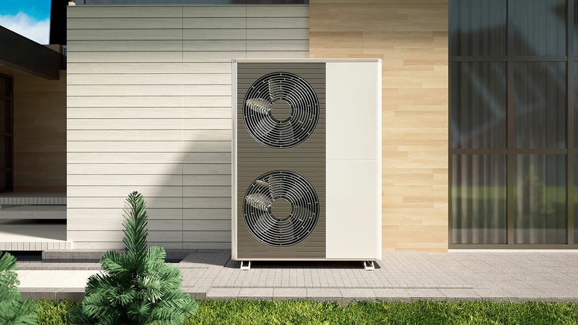 A heat pump is situated outside a modern home with wood siding. The unit is white with two fans and is positioned next to a window. Lush green grass covers the ground in front of the home.